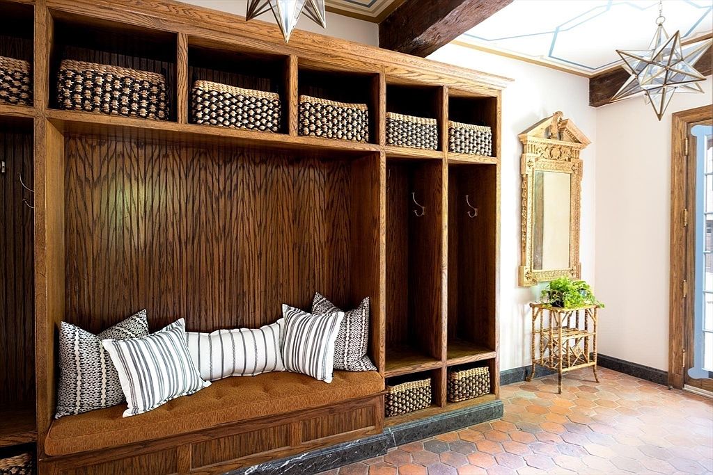 This elegant mudroom entryway features custom floor-to-ceiling dark wood cabinetry with integrated bench seating and woven storage baskets. The space is accented by a decorative gold-framed mirror, a rustic rattan side table with greenery, and a unique star-shaped pendant light fixture. The warm, earthy tones and terracotta tile flooring create a sophisticated and welcoming atmosphere.