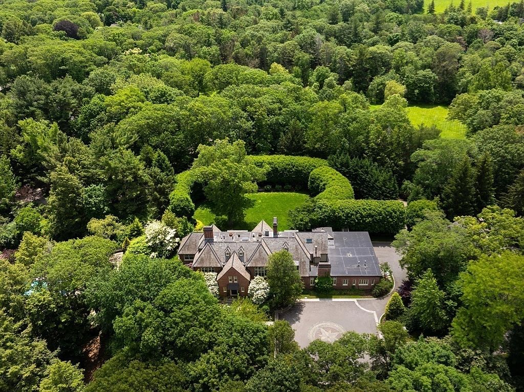 This high-angle aerial view captures a sprawling, traditional brick estate nestled within a dense, lush forest canopy. The property features a circular driveway with a decorative emblem, a manicured lawn enclosed by a tall, perfectly shaped hedge, and a multi-gabled roofline that adds architectural character. The perspective emphasizes the home's secluded, park-like setting and its grand scale within the surrounding landscape.