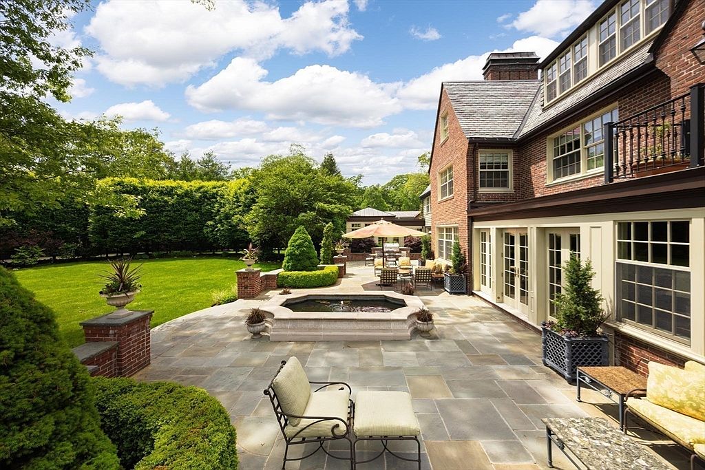 This elegant rear patio features high-quality stone paving, a central octagonal fountain, and comfortable outdoor seating arrangements. The space is framed by a classic brick home with large glass doors, lush manicured hedges, and a sprawling green lawn, creating a serene and sophisticated atmosphere. The perspective is a wide, eye-level shot that captures the seamless transition between the refined indoor living space and the expansive, well-maintained garden.