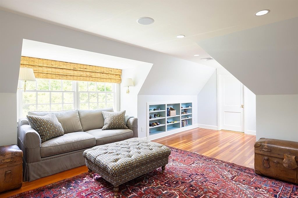 This cozy attic living space features sloped ceilings, warm hardwood flooring, and a large window dressed with bamboo shades. The room is furnished with a comfortable striped sofa, a patterned ottoman, and built-in shelving, creating a welcoming and functional retreat. The perspective is a wide-angle shot that captures the inviting atmosphere and architectural character of the space.