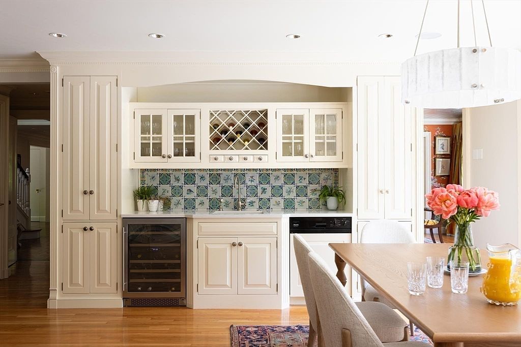 This elegant kitchen features a classic, light-toned aesthetic with cream-colored cabinetry, a decorative patterned tile backsplash, and a built-in wine refrigerator. A wooden dining table with upholstered chairs sits in the foreground, creating a warm and inviting atmosphere perfect for entertaining. The perspective is a straight-on, eye-level shot that highlights the seamless integration of the wet bar area with the adjacent dining space.