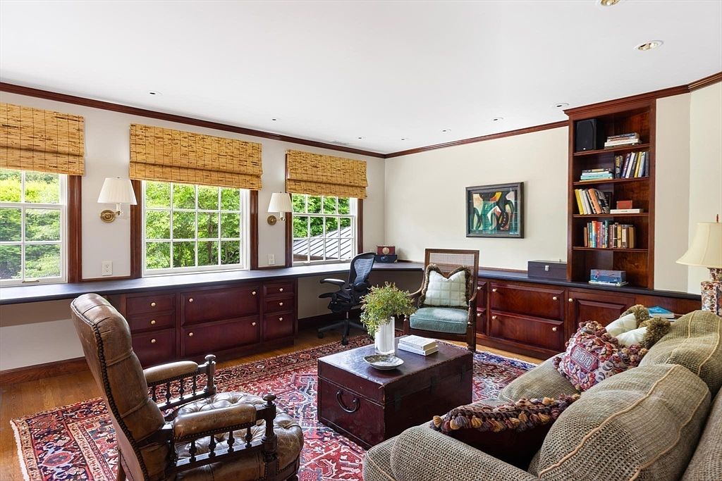 This sophisticated home office features rich, dark wood built-in cabinetry and a matching desk that spans the length of the window wall. The room is anchored by a large, ornate Persian-style rug, a leather armchair, and a comfortable sofa, creating a warm and scholarly atmosphere. Natural light streams through three large windows adorned with bamboo shades, highlighting the classic decor and the well-stocked bookshelf.