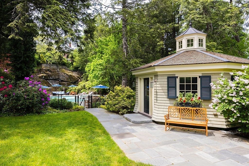 This serene backyard scene features a charming, light-colored gazebo with a cupola, set beside a stone-paved walkway and a lush green lawn. A wooden bench sits invitingly in front of the gazebo, while a swimming pool with blue umbrellas is visible in the background, nestled against a natural rock formation. The setting is framed by mature trees and vibrant flowering bushes, creating a peaceful and private outdoor retreat.