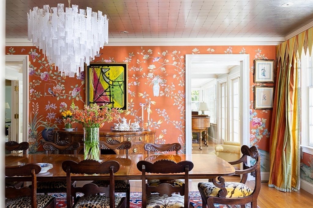 This vibrant dining room features bold orange floral wallpaper, a polished wooden dining table, and ornate carved wooden chairs with patterned upholstery. A striking, modern white glass-shard chandelier hangs above the table, while a large abstract painting adds a contemporary touch to the traditional decor. The room is framed by elegant doorways and floor-to-ceiling curtains, creating a sophisticated and eclectic atmosphere.