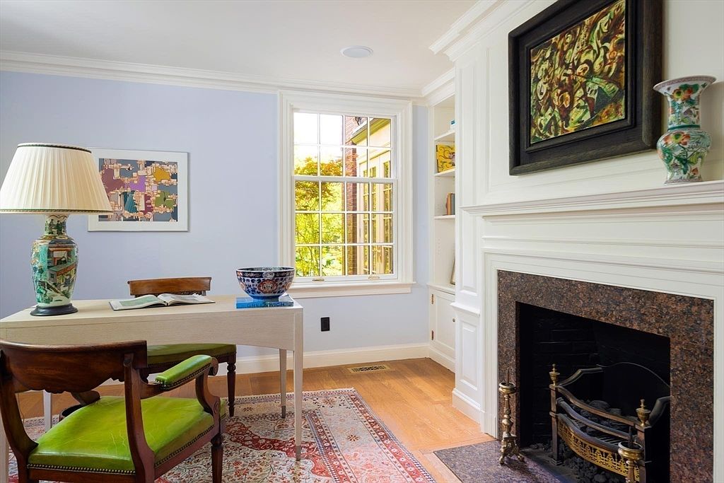 This elegant home office features a classic desk paired with a vibrant green leather chair, set against a backdrop of soft blue walls and a traditional fireplace with a dark granite surround. A large window allows natural light to fill the room, highlighting the hardwood floors and built-in shelving, while a decorative painting above the mantel adds a sophisticated touch. The overall aesthetic is a refined blend of traditional charm and functional workspace design.