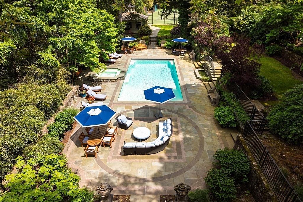 This high-angle aerial view captures a luxurious, secluded backyard oasis featuring a rectangular swimming pool and an adjacent hot tub set within a spacious stone patio. The area is elegantly furnished with multiple outdoor seating arrangements, including a curved sectional sofa and a dining set, all shaded by blue umbrellas. Surrounded by lush, mature greenery and trees, the space offers a private, resort-like atmosphere perfect for relaxation and outdoor entertaining.