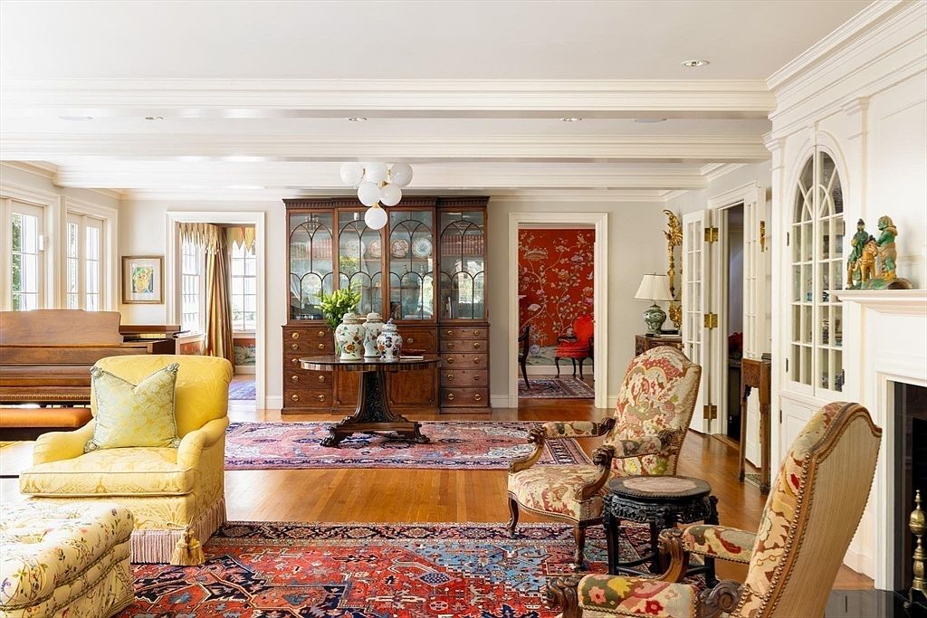 This elegant, traditional living room features a warm color palette with rich hardwood floors and intricate Persian-style area rugs. The space is anchored by a large wooden hutch, a yellow upholstered armchair, and patterned accent chairs, all illuminated by a modern globe chandelier. The room offers a sophisticated, classic aesthetic with a view into an adjacent room featuring vibrant red wallpaper, creating a sense of depth and luxury.