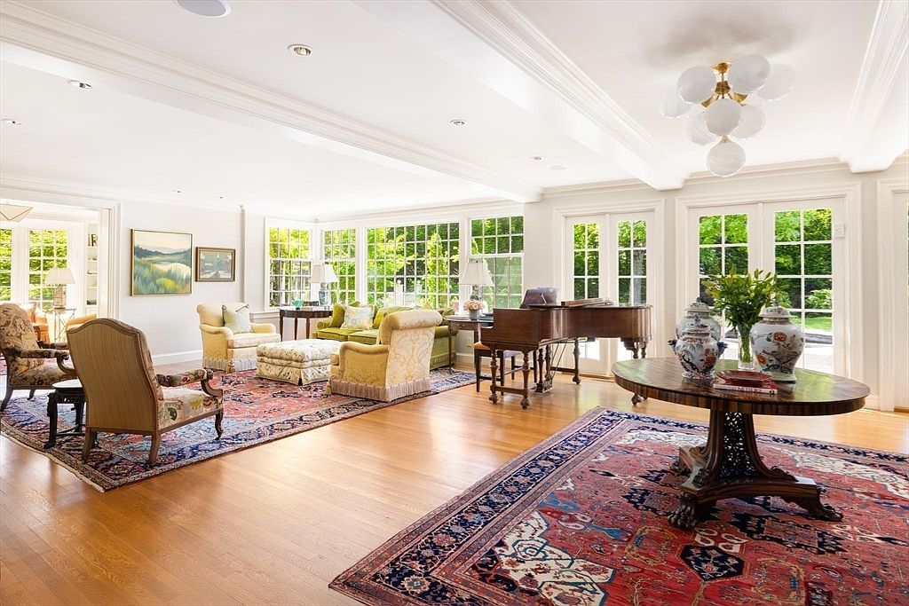 This expansive and elegant living room features polished hardwood floors, high ceilings with recessed lighting, and a grand piano, creating a sophisticated atmosphere. The space is bathed in natural light from large windows and French doors that overlook the lush greenery outside, while traditional furniture and ornate area rugs add warmth and character. The open-concept layout seamlessly blends multiple seating areas, making it an ideal space for both relaxation and formal entertaining.