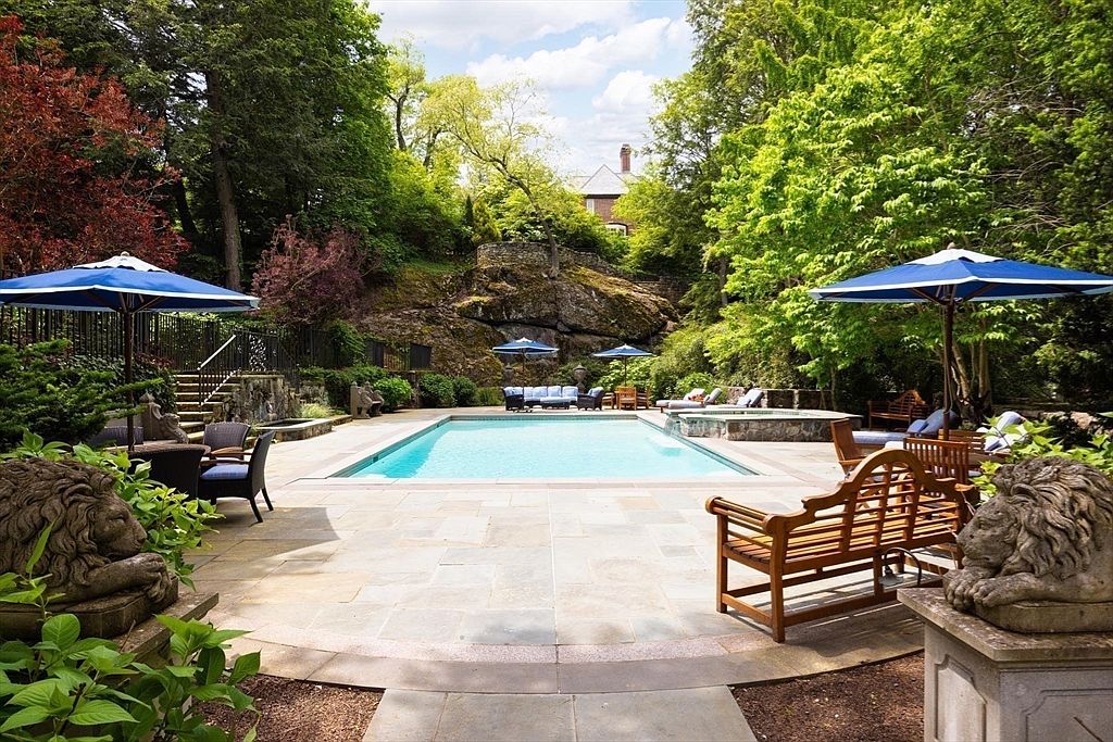This luxurious backyard features a pristine rectangular swimming pool set against a dramatic, natural rock cliff backdrop. The expansive stone patio is furnished with multiple seating areas, blue umbrellas, and decorative stone lion statues, creating an elegant and serene outdoor retreat. The lush surrounding greenery and mature trees provide a sense of privacy and seclusion, perfect for high-end entertaining.