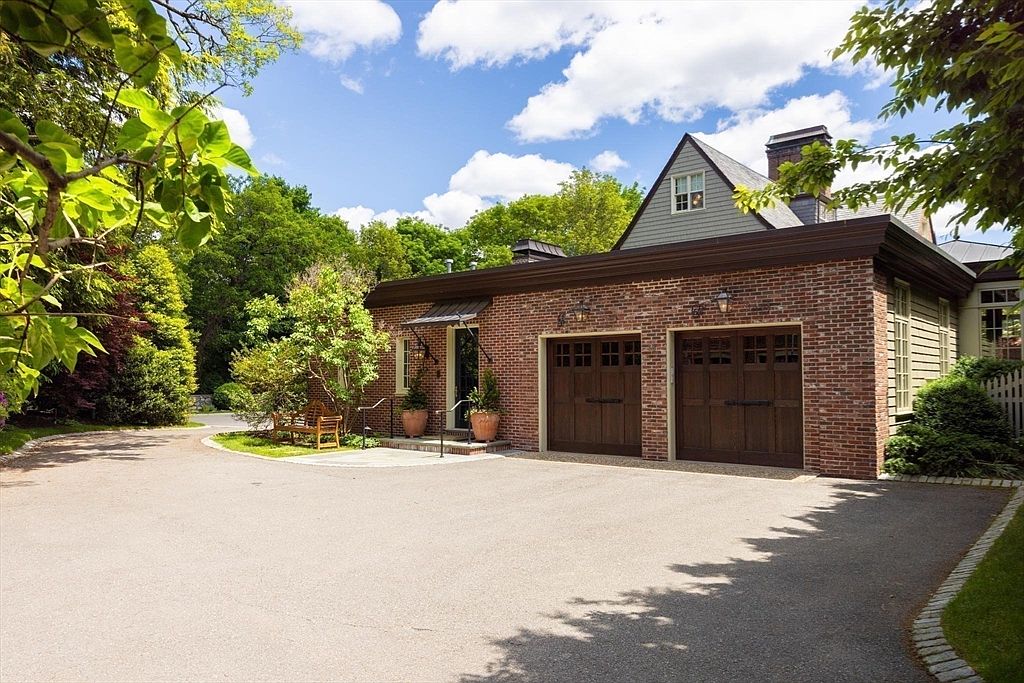 This image showcases a charming, traditional-style brick garage with two dark wood carriage-style doors and a matching side entry door. The structure features a classic gable roofline with a dormer window above, set against a backdrop of lush green trees and a clear blue sky. A paved driveway leads up to the garage, complemented by a small landscaped area with a wooden bench and potted plants, creating a welcoming and well-maintained curb appeal.