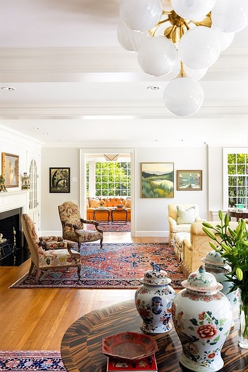 This elegant living room features a sophisticated blend of traditional and transitional styles, anchored by a vibrant, intricately patterned area rug. The space is illuminated by a striking, large-scale globe chandelier and flows seamlessly into a sun-drenched conservatory through a wide doorway. In the foreground, a polished wood table displays ornate ceramic ginger jars, while comfortable seating arrangements create a warm and inviting atmosphere perfect for entertaining.