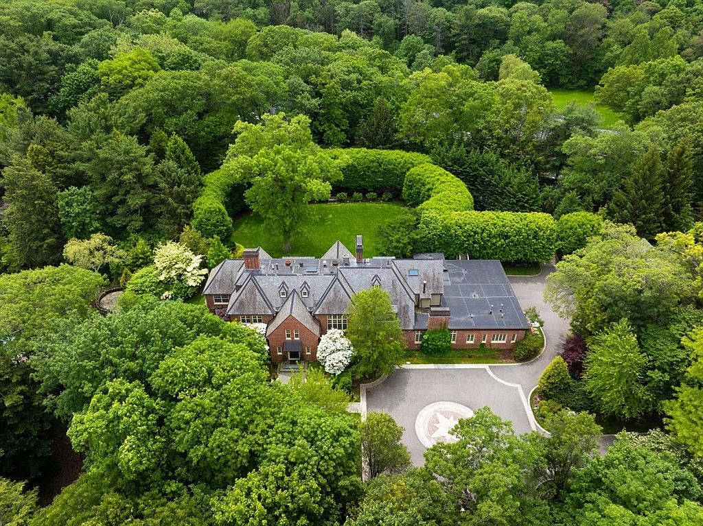 This high-angle aerial shot captures a sprawling, traditional brick estate nestled within a dense, lush forest canopy. The property features a grand circular driveway with a decorative stone inlay, a meticulously manicured lawn enclosed by tall, sculpted hedges, and a complex multi-gabled roofline. The perspective emphasizes the home's secluded, park-like setting and its impressive architectural scale.