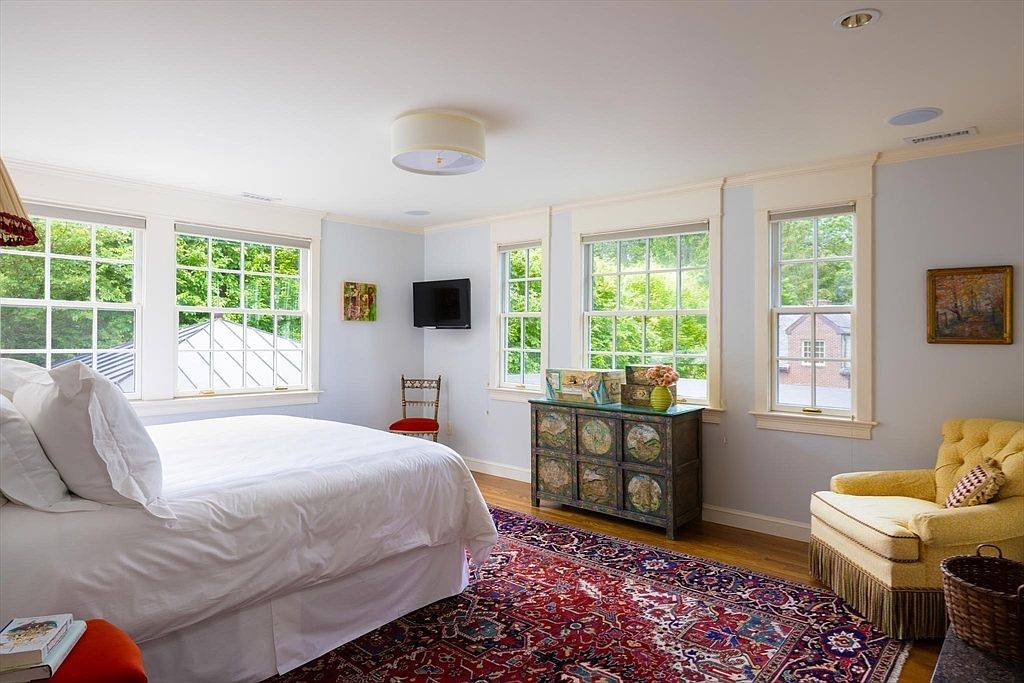 This bright and airy primary bedroom features a large, plush bed with white linens, a vibrant red patterned area rug, and a classic yellow upholstered armchair. The room is filled with natural light from multiple windows, highlighting the light blue walls and a unique, hand-painted wooden chest of drawers. The overall aesthetic is traditional and cozy, offering a serene and inviting atmosphere.