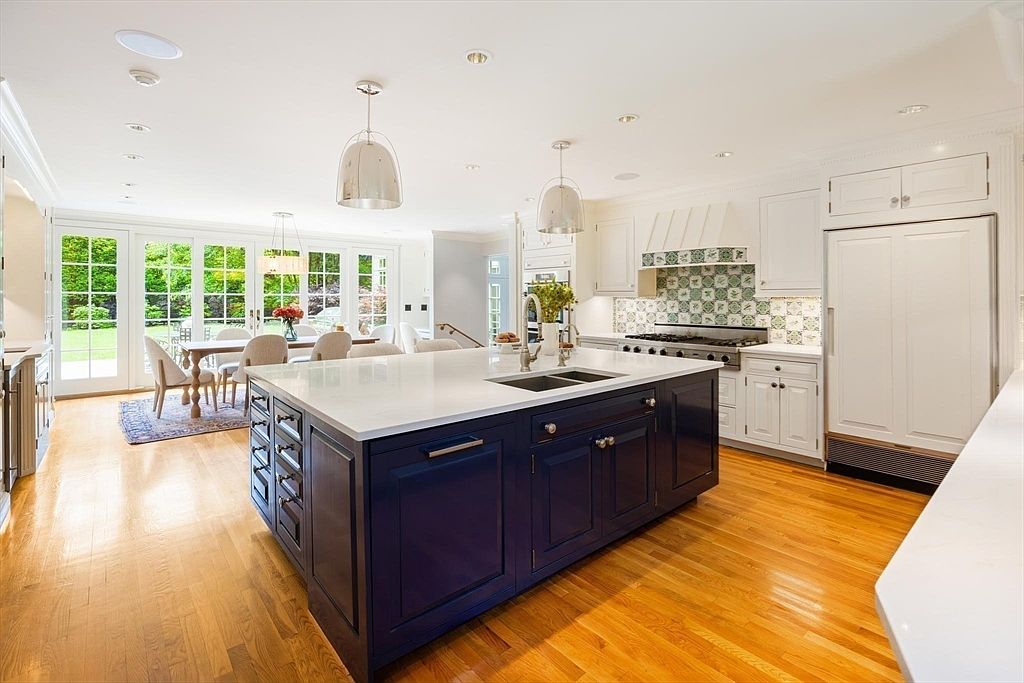 This bright and spacious kitchen features a striking navy blue center island with a white quartz countertop, contrasting beautifully against the surrounding white cabinetry and hardwood floors. The space is illuminated by elegant pendant lighting and recessed ceiling lights, while a large dining area is visible through the open floor plan, overlooking a lush green backyard. The overall aesthetic is a sophisticated blend of classic and modern design, perfect for both cooking and entertaining.