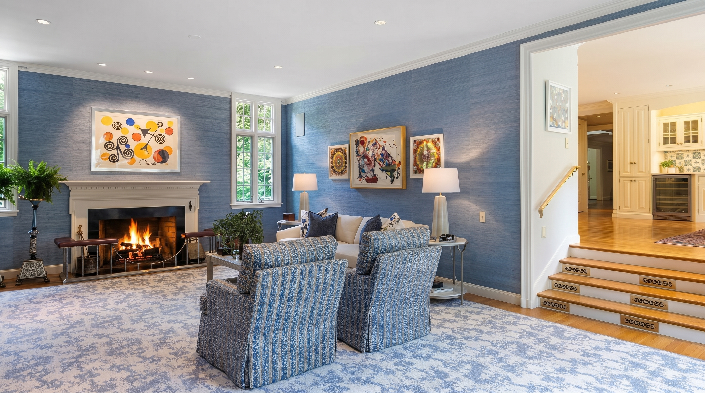 This elegant living room features textured blue wallpaper, a classic fireplace with a decorative mantel, and a plush patterned carpet. The space is furnished with two comfortable armchairs and a sofa, accented by abstract artwork and warm table lamps. A set of wooden stairs leads up to an adjacent room, creating an inviting and sophisticated atmosphere.