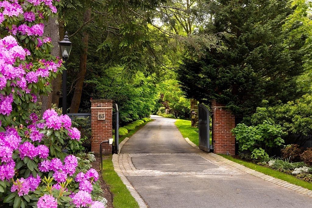 This inviting entryway features a private, tree-lined driveway framed by classic brick pillars and open wrought-iron gates. A vibrant, blooming rhododendron bush in the foreground adds a splash of color, while the paved driveway leads the eye toward a lush, secluded landscape. The scene conveys a sense of privacy, elegance, and a well-maintained estate entrance.