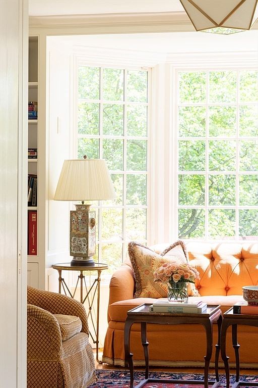 This inviting living room features a vibrant orange tufted sofa accented with patterned throw pillows and a bouquet of fresh roses. A classic bay window floods the space with natural light, highlighting a decorative table lamp with an Asian-inspired base and a patterned armchair. The room exudes a warm, traditional elegance, perfect for a cozy reading nook or a sophisticated sitting area.
