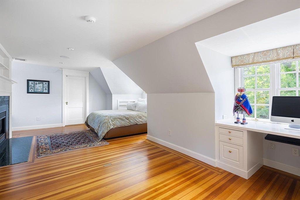 This spacious, light-filled bedroom features a unique attic-style sloped ceiling and warm, polished hardwood floors. The room is thoughtfully laid out with a cozy bed area and a built-in desk workspace nestled under a window, creating a versatile living environment. A fireplace on the left and a classic rug add a touch of traditional charm to this bright and airy space.