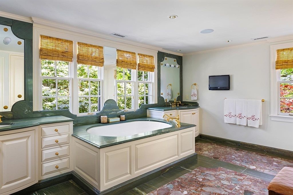 This elegant primary bathroom features a central soaking tub framed by custom green marble countertops and white cabinetry. The space is bathed in natural light from large windows adorned with bamboo shades, creating a serene and luxurious atmosphere. A wall-mounted television and plush towels add modern convenience to the classic, sophisticated design.