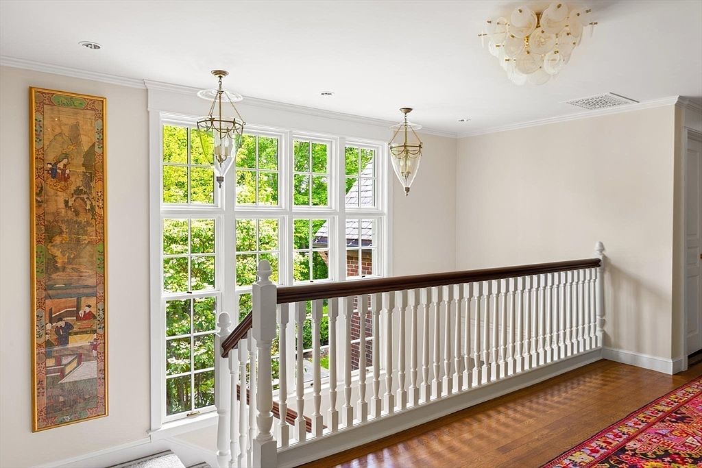 This elegant upper-level landing features polished hardwood floors, a classic white wooden banister, and a vibrant, patterned runner rug. The space is illuminated by a sophisticated multi-tiered chandelier and two hanging glass lanterns, while a tall, traditional Asian-style scroll painting adds a unique decorative touch. Large, multi-pane windows provide ample natural light and a view of the lush greenery outside, creating a bright and welcoming atmosphere.