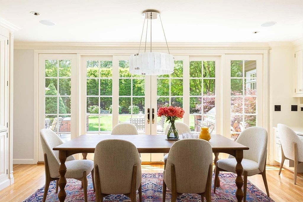 This elegant dining room features a warm wooden table surrounded by six upholstered chairs, set atop a vibrant, patterned area rug. A modern, cylindrical chandelier hangs above, while large glass-paned French doors provide an abundance of natural light and a scenic view of the lush backyard. The space is accented by a vase of fresh flowers and a pitcher, creating a welcoming and sophisticated atmosphere perfect for hosting.
