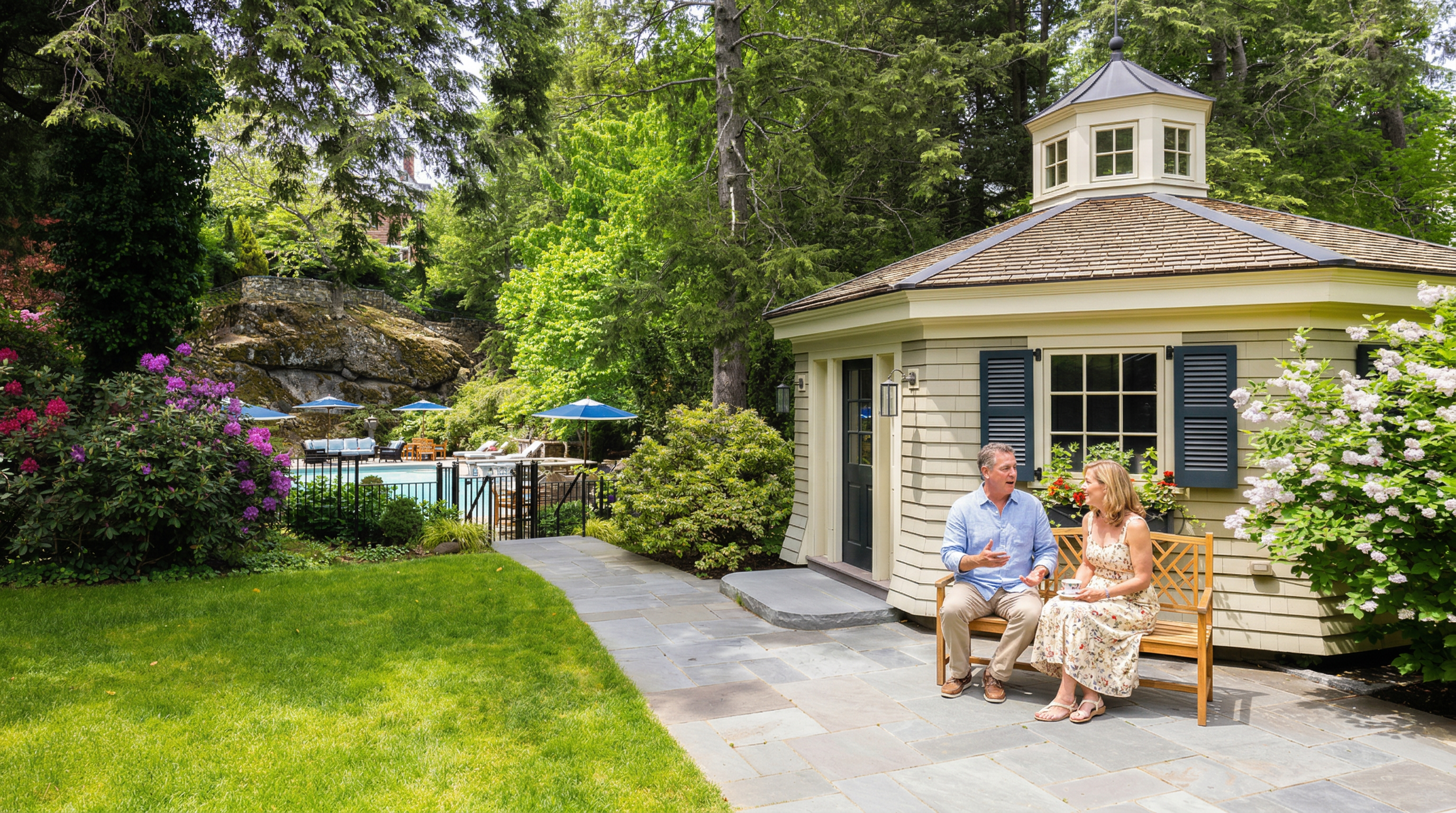 This serene backyard scene features a charming, light-colored gazebo with a cupola, set beside a stone-paved walkway and a lush green lawn. A wooden bench sits invitingly in front of the gazebo, while a swimming pool with blue umbrellas is visible in the background, nestled against a natural rock formation. The setting is framed by mature trees and vibrant flowering bushes, creating a peaceful and private outdoor retreat.