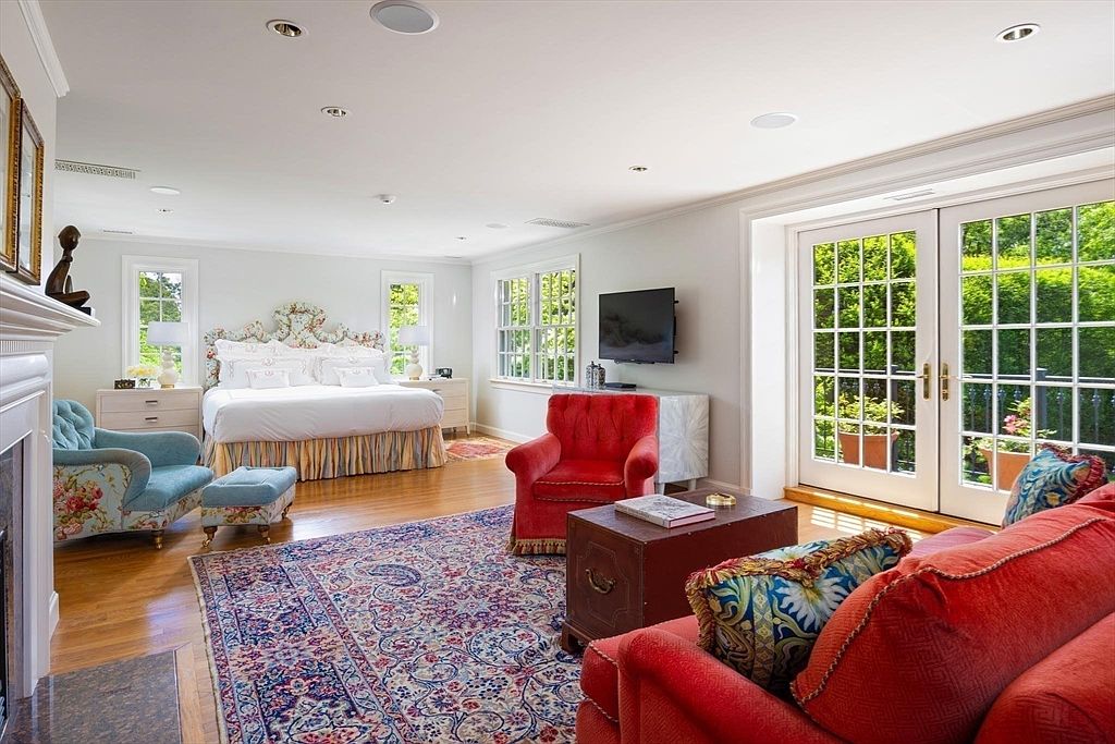 This spacious primary bedroom features a classic, elegant design with a large bed as the focal point, complemented by a vibrant, traditional area rug and a cozy seating area. The room is bathed in natural light from the French doors that open to an outdoor space, while a fireplace and television provide comfort and entertainment. The combination of soft neutral walls and bold, colorful furniture creates a sophisticated and inviting atmosphere.