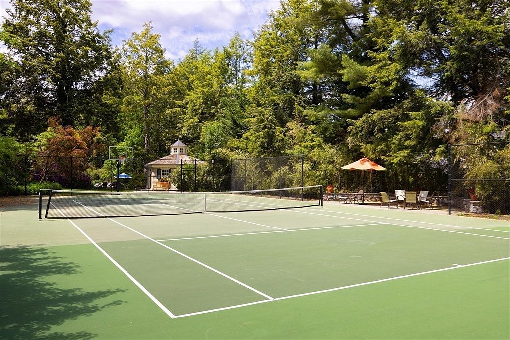 This image showcases a well-maintained private tennis court nestled within a lush, wooded landscape. The scene includes a small gazebo in the background and a patio area with outdoor seating shaded by an umbrella, creating a serene and recreational atmosphere. The perspective is a wide, eye-level shot that emphasizes the expansive outdoor space and the peaceful, nature-surrounded setting.