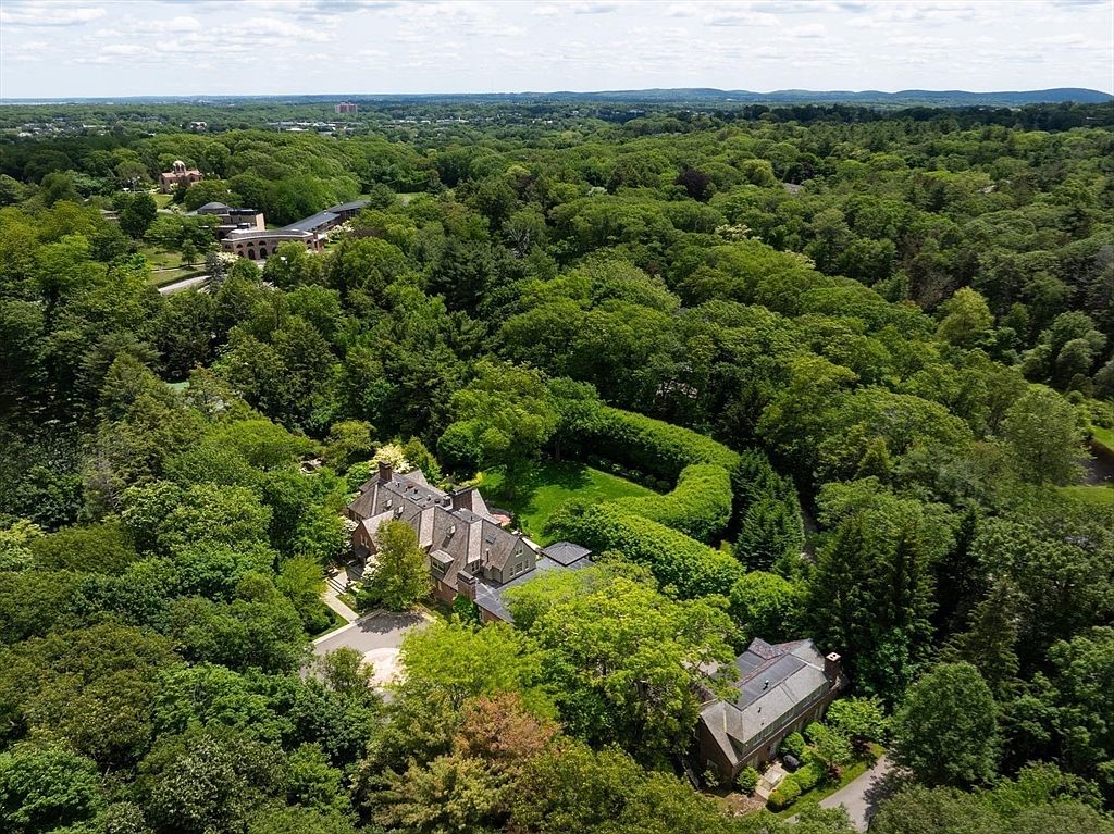 This high-angle aerial view captures a sprawling, secluded estate nestled within a dense, lush forest canopy. The property features a large, multi-gabled traditional home with a circular driveway and a distinct, manicured hedge-lined garden area. The perspective emphasizes the expansive, private nature of the grounds and the serene, wooded surroundings.