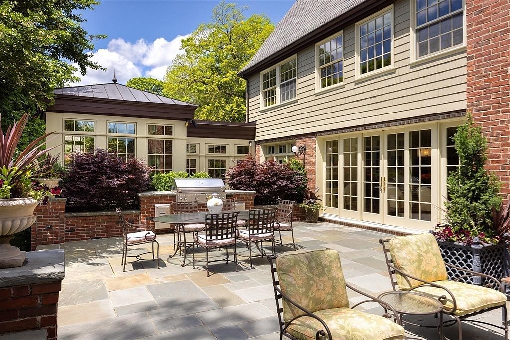 This elegant stone patio features a built-in outdoor kitchen with a stainless steel grill, perfect for al fresco dining and entertaining. The space is furnished with a dining set and comfortable lounge chairs, all set against the backdrop of a charming brick and siding home with large glass doors. Lush greenery and mature trees surround the area, creating a serene and private outdoor living environment.
