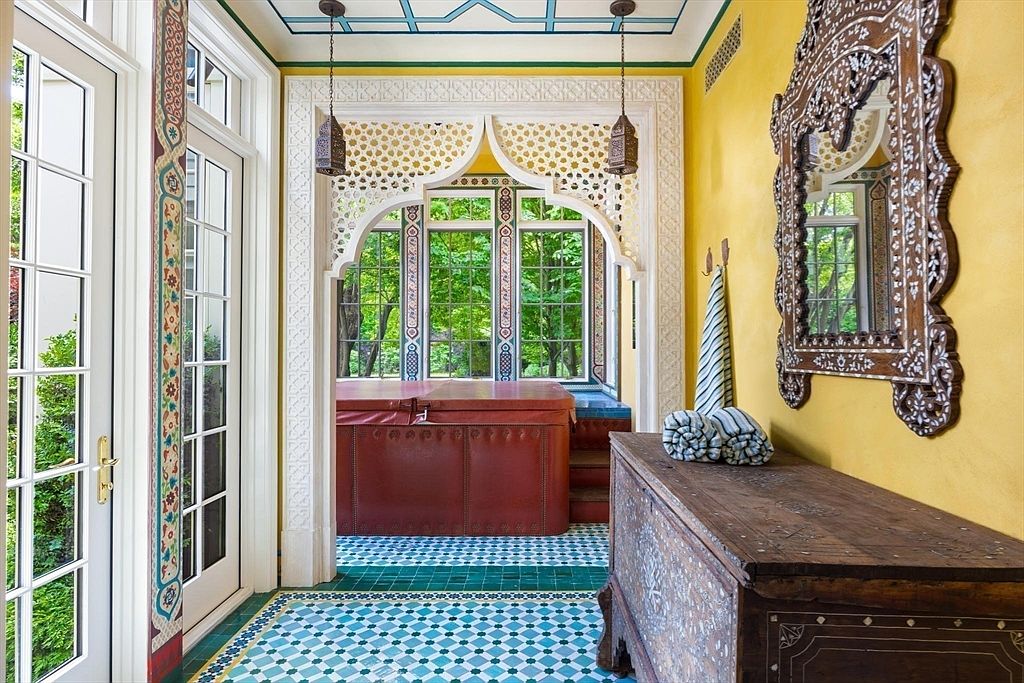 This unique sunroom features a striking Moroccan-inspired aesthetic, highlighted by intricate white lattice woodwork, vibrant geometric floor tiles, and a deep red hot tub as the central focal point. The room is framed by bright yellow walls and an ornate, inlaid wooden chest, creating a warm and exotic atmosphere. Large windows provide a seamless connection to the lush greenery outside, enhancing the tranquil and luxurious feel of the space.