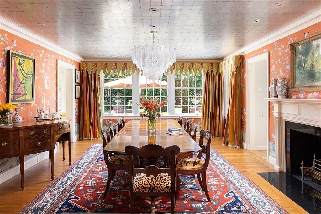 This elegant dining room features a long wooden table surrounded by chairs with animal-print upholstery, set atop a vibrant, traditional-style area rug. The space is defined by bold orange floral wallpaper, a unique metallic grid-patterned ceiling, and a grand crystal chandelier that serves as a focal point. A fireplace with a mantel and a classic sideboard complete the sophisticated, formal aesthetic of the room.