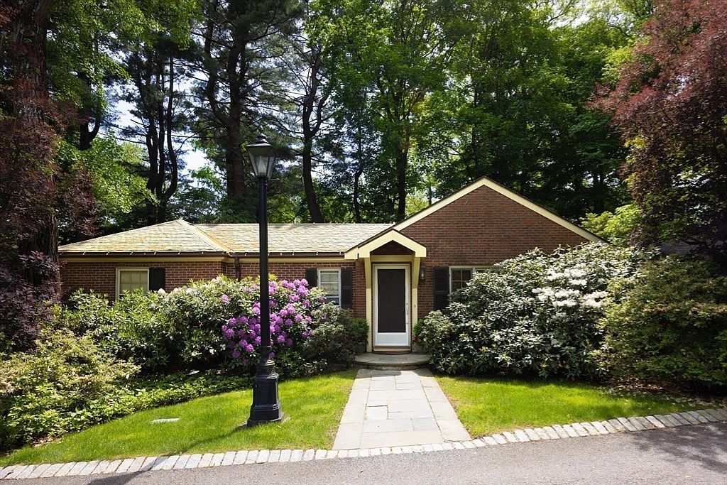 This charming single-story brick home features a classic architectural style nestled within a lush, wooded landscape. A stone walkway leads directly to the front entryway, which is framed by a small gable roof and surrounded by vibrant, blooming rhododendron bushes. The scene is captured from a low-angle perspective, emphasizing the home's inviting curb appeal and serene, natural setting.