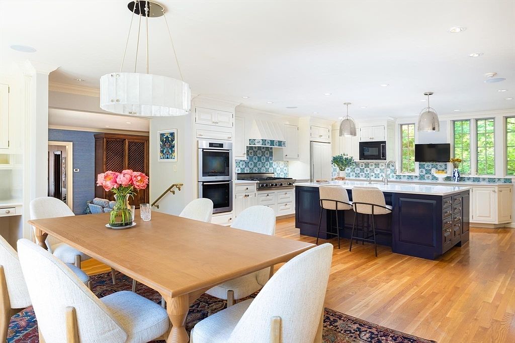 This bright and spacious open-concept kitchen features a large navy blue center island with bar seating, complemented by white cabinetry and a patterned tile backsplash. A wooden dining table sits in the foreground, creating a seamless transition between the dining and cooking areas. The room is illuminated by elegant pendant lighting and natural light from large windows, offering a sophisticated and inviting atmosphere.