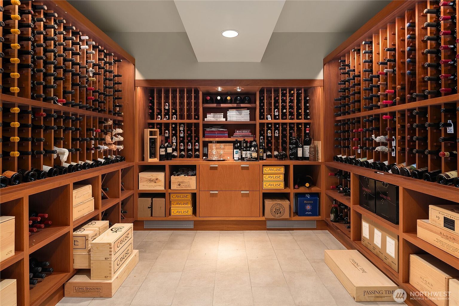 This sophisticated, custom-built wine cellar features extensive floor-to-ceiling wooden racking designed to showcase a large collection of bottled wines. The room is organized with dedicated shelving for individual bottles, wooden storage crates on the floor, and a central display area with drawers and counter space. The warm wood tones, recessed lighting, and tiled flooring create an elegant and climate-controlled atmosphere perfect for wine enthusiasts.