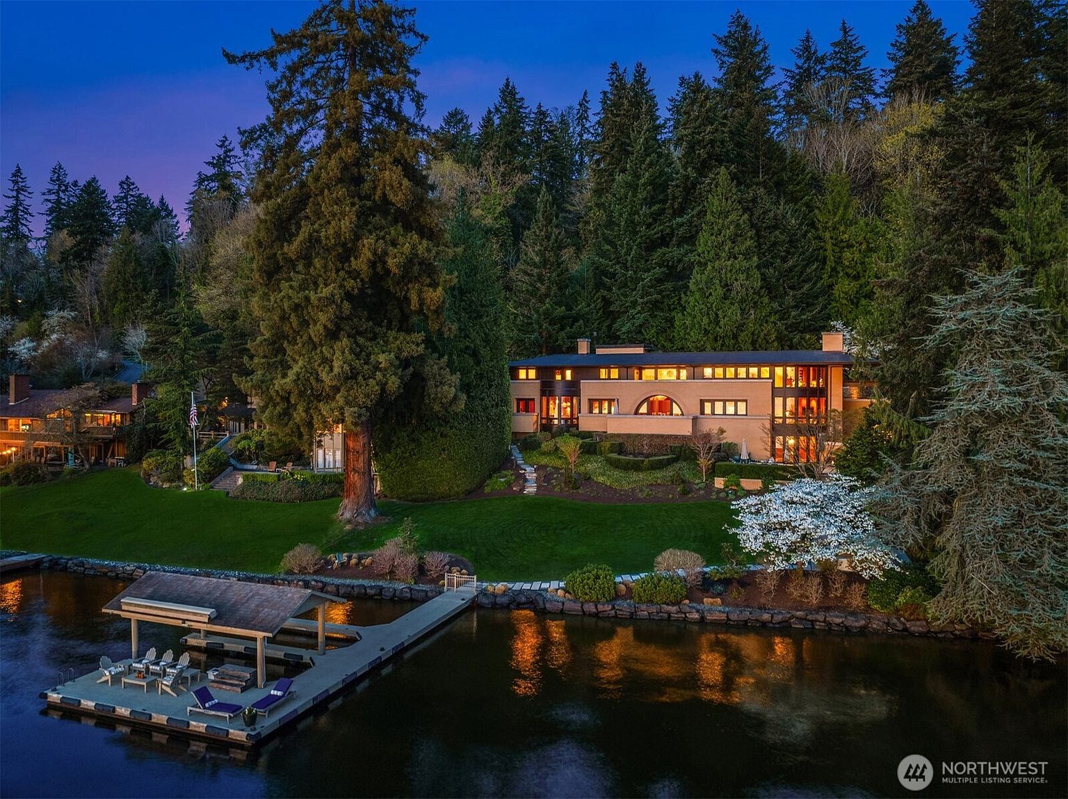 This stunning aerial view captures a luxurious waterfront estate nestled against a lush, dense forest backdrop at twilight. The multi-level home features warm, inviting interior lighting that contrasts beautifully with the deep blue evening sky, while a private dock with a covered seating area extends into the calm water. The meticulously landscaped lawn and mature trees create a serene, private atmosphere, highlighting the property's prime location and architectural elegance.