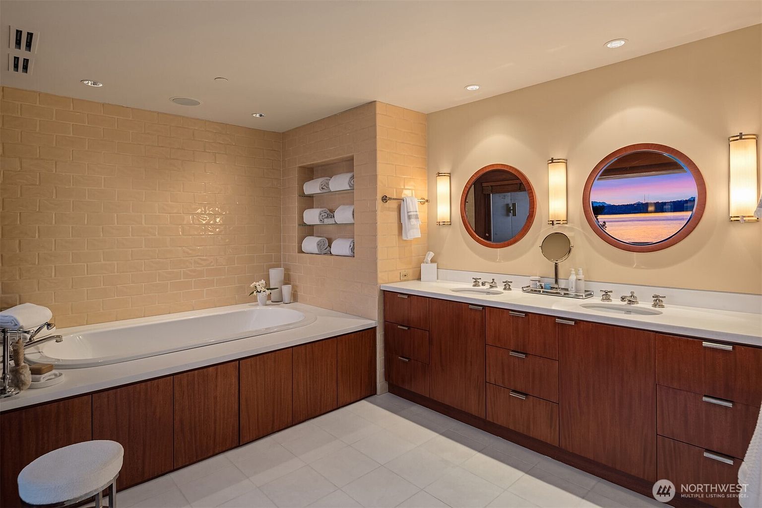 This luxurious primary bathroom features a sleek, modern design with a deep soaking tub set against a textured, light-colored brick tile wall. The vanity area includes a long wooden cabinet with dual sinks, complemented by circular mirrors that reflect a serene waterfront view. The space is well-lit with elegant wall sconces, creating a warm and sophisticated atmosphere.