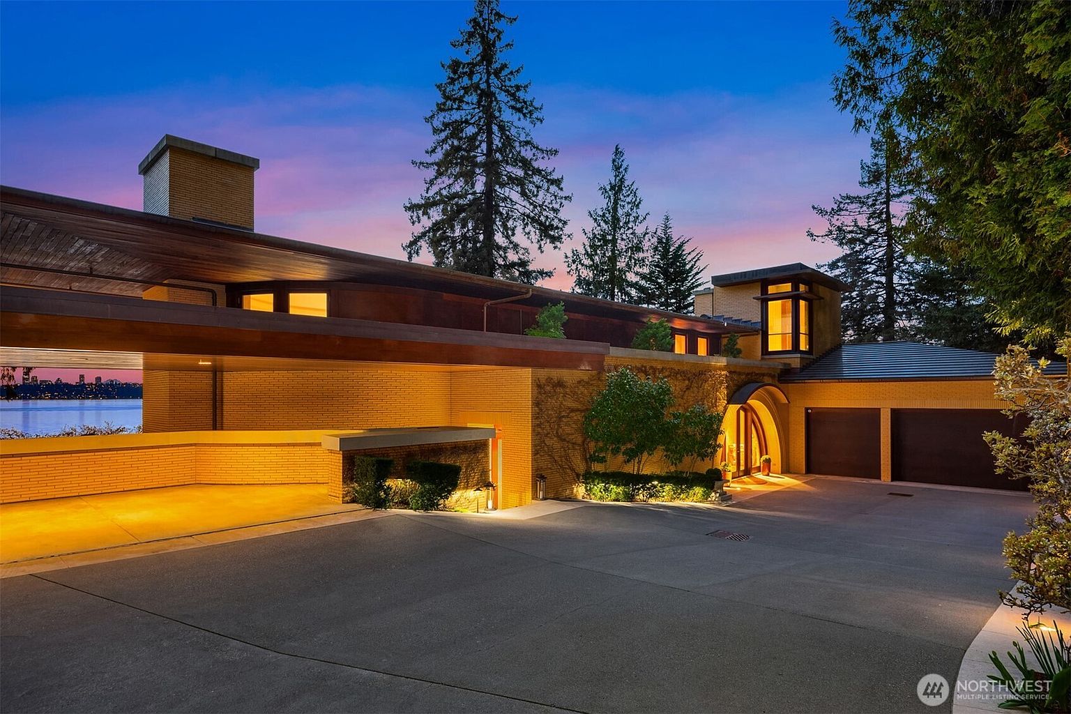 This striking mid-century modern residence features a low-slung, horizontal profile with warm yellow brick masonry and expansive wood-paneled overhangs. The driveway leads to a multi-car garage and a dramatic, arched entryway, all illuminated by soft, inviting landscape lighting against a vibrant twilight sky. The property offers a unique perspective with a view of the water and city skyline visible through the open carport structure.
