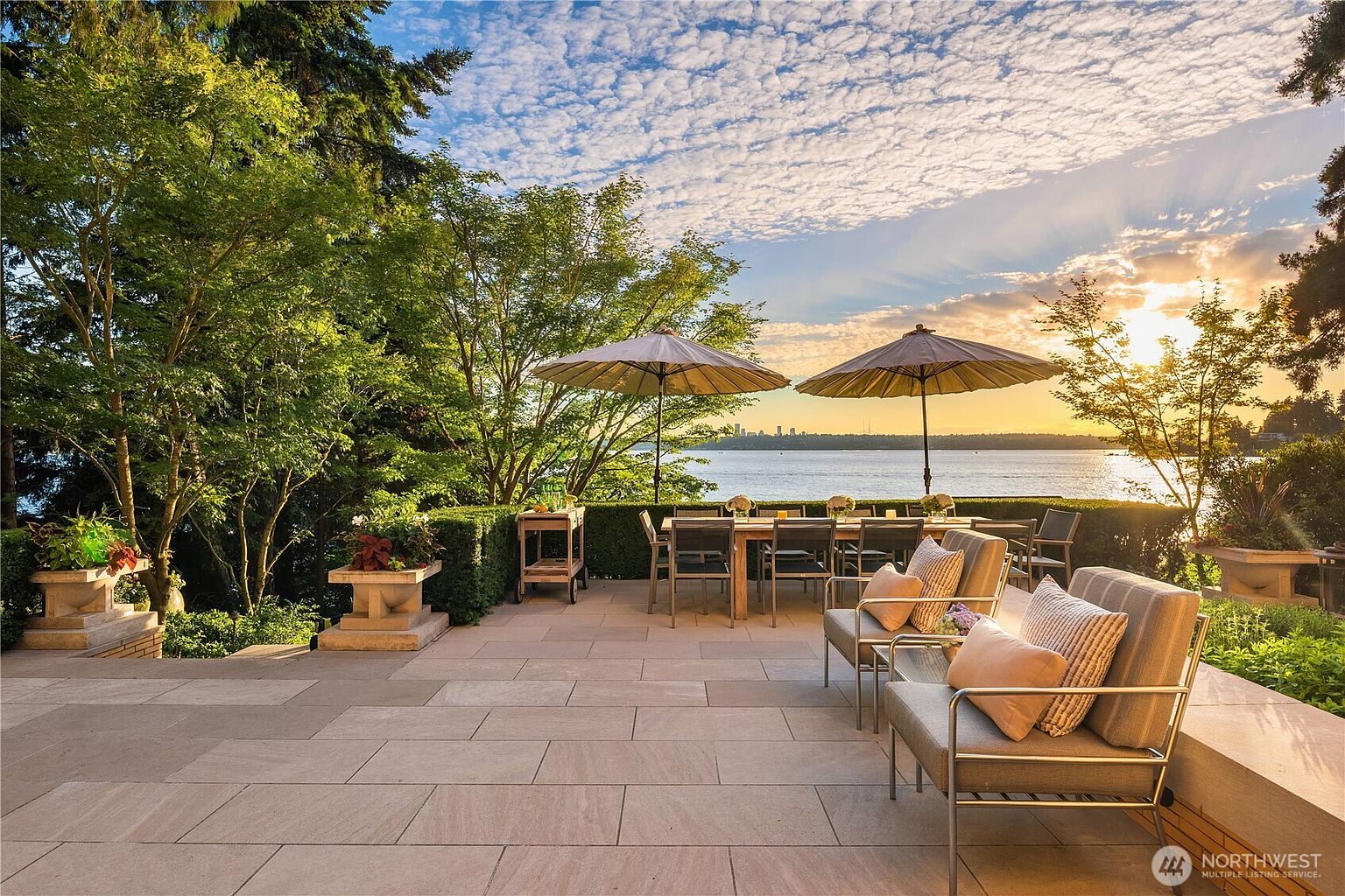 This stunning stone-paved patio offers a luxurious outdoor living space with a dining area and comfortable lounge seating, all overlooking a serene waterfront view. The scene is framed by lush green trees and two large umbrellas, creating a perfect setting for sunset relaxation with a distant city skyline visible on the horizon. The overall atmosphere is one of sophisticated tranquility and high-end waterfront living.