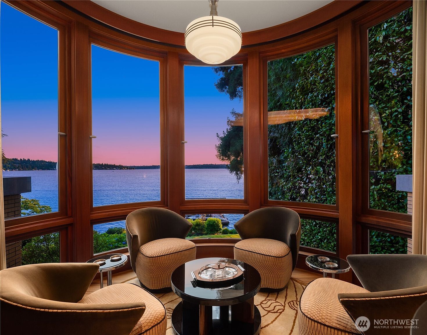 This elegant seating area features a curved wall of floor-to-ceiling windows that frame a breathtaking sunset view over the water. The room is furnished with four sophisticated, mid-century modern-inspired armchairs arranged around a sleek, circular black coffee table, all set atop a patterned area rug. A classic pendant light fixture hangs from the ceiling, enhancing the warm, luxurious atmosphere of this serene waterfront retreat.