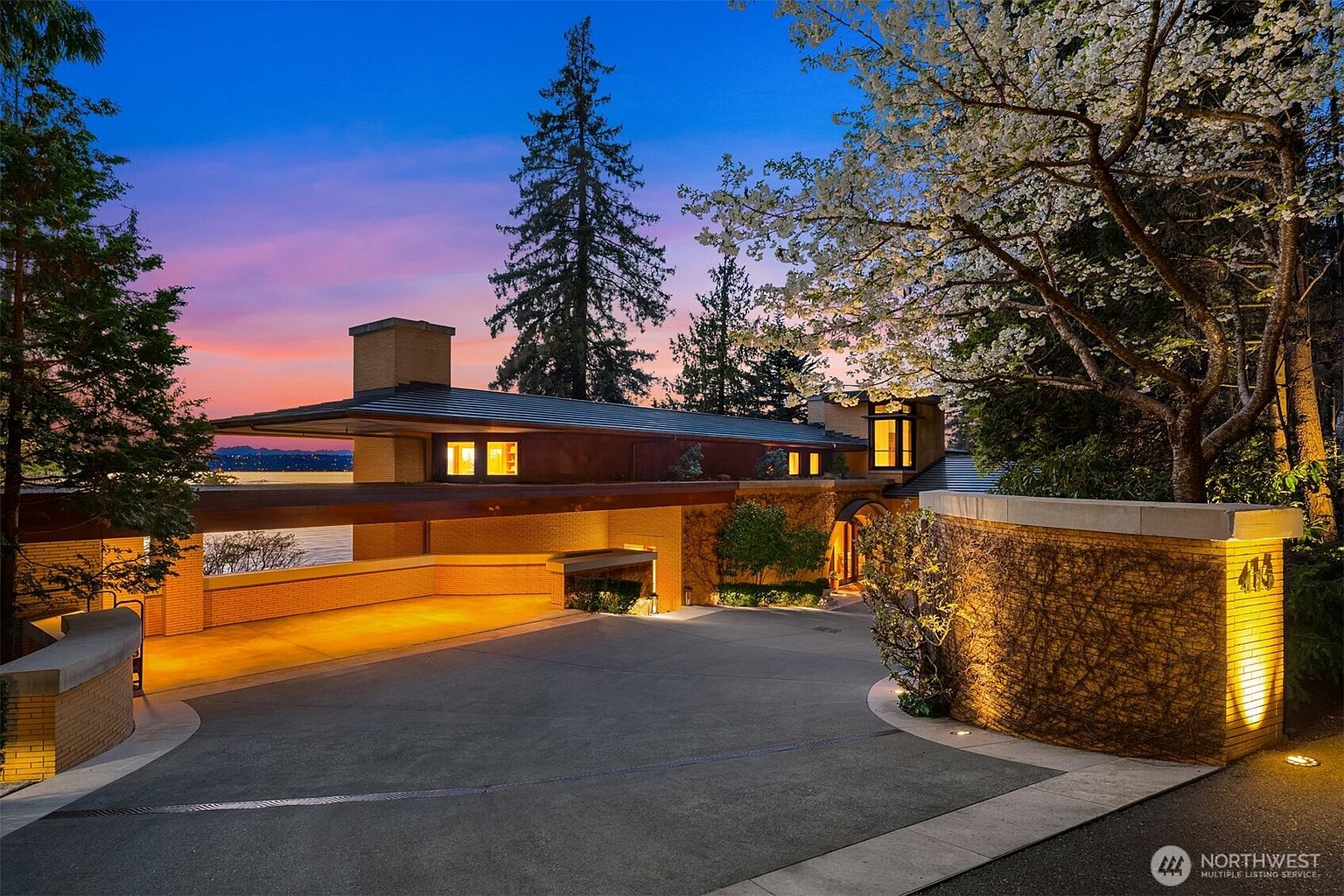 This striking mid-century modern residence is captured at dusk, showcasing its low-slung, horizontal architecture and warm, integrated lighting. The property features a prominent brick driveway leading to a covered carport, with a beautifully illuminated stone wall in the foreground and a blooming tree adding a soft, organic contrast to the clean lines. The composition emphasizes the home's seamless blend with the natural landscape and its serene waterfront setting.