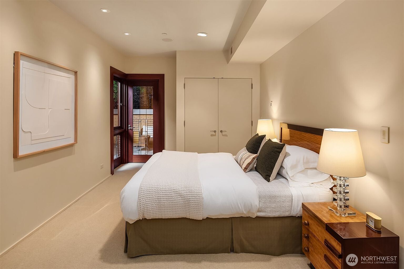 This primary bedroom features a neutral color palette, plush carpeting, and a large bed centered against a wood-accented headboard. A glass-paned door provides direct access to an outdoor patio, while recessed lighting and bedside lamps create a warm, inviting atmosphere. The room is clean and minimalist, offering a serene and comfortable living space.