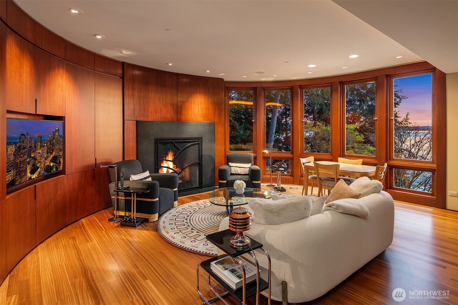 This sophisticated living room features warm wood-paneled walls, a cozy fireplace, and a curved layout that emphasizes the expansive floor-to-ceiling windows overlooking a scenic waterfront. The space is furnished with a plush white sofa, dark accent chairs, and a circular patterned rug, creating a luxurious and inviting atmosphere. The cinematic perspective captures the seamless blend of high-end interior design and the natural beauty of the outdoor landscape.