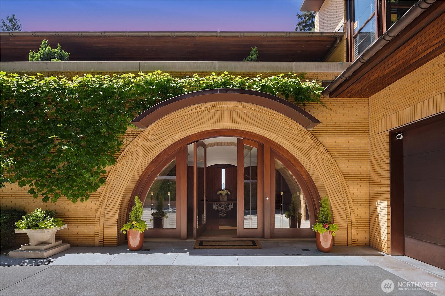 This striking entryway features a prominent, oversized arched wooden door set within a decorative brick facade, framed by lush climbing ivy. The symmetrical composition includes two potted plants flanking the entrance, creating a welcoming and sophisticated architectural focal point. The perspective is a direct, eye-level shot that emphasizes the unique craftsmanship and grand scale of the home's front entrance.
