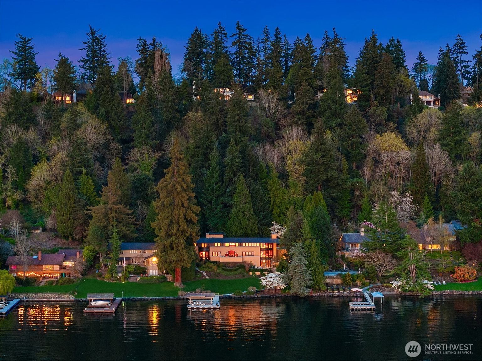This stunning aerial view captures a luxurious waterfront estate nestled against a lush, forested hillside at twilight. The property features multiple multi-level homes with warm, inviting interior lighting, expansive green lawns, and private boat docks extending into the calm lake. The scene evokes a sense of serene, high-end living, perfectly blending architectural elegance with the natural beauty of the surrounding woodland.