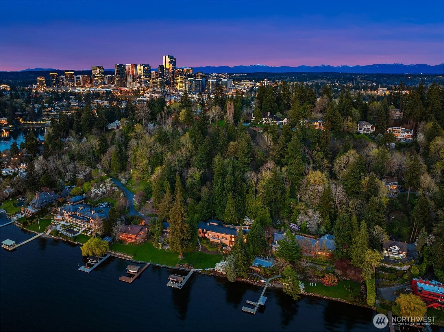 This stunning aerial view captures a luxurious waterfront estate nestled within a lush, forested landscape at twilight. The property features multiple large homes with private docks extending into the calm water, all set against the backdrop of a vibrant city skyline and distant mountain ranges. The perspective provides a cinematic sense of scale, highlighting the peaceful seclusion of the homes while emphasizing their proximity to urban amenities.