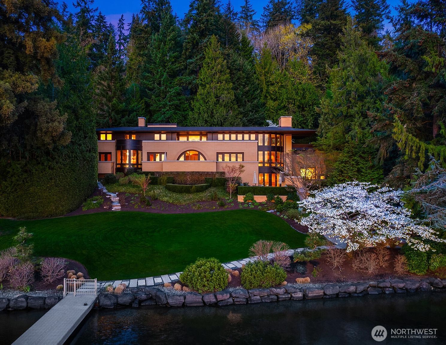 This stunning waterfront estate showcases a sophisticated architectural design reminiscent of Frank Lloyd Wright's Prairie style, nestled against a lush, dense forest backdrop. The multi-level home features warm-toned brick, expansive windows, and a prominent arched window, all illuminated by a soft, inviting glow at dusk. A manicured lawn leads down to a private dock on the water, framed by a blooming white tree, creating a serene and luxurious atmosphere.