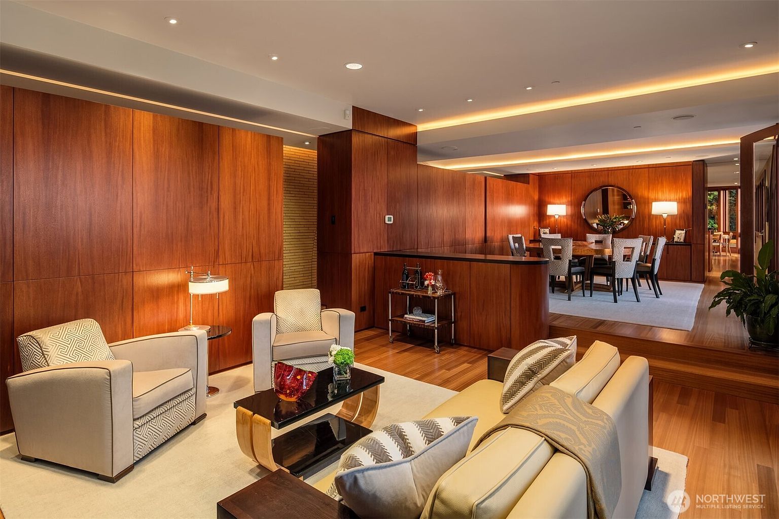 This sophisticated living room features rich wood-paneled walls and a warm, inviting atmosphere, seamlessly transitioning into an elevated dining area. The space is furnished with plush, neutral-toned seating and a sleek coffee table, all illuminated by recessed lighting and elegant lamps. The open-concept layout and high-end finishes create a luxurious, cinematic feel perfect for upscale entertaining.