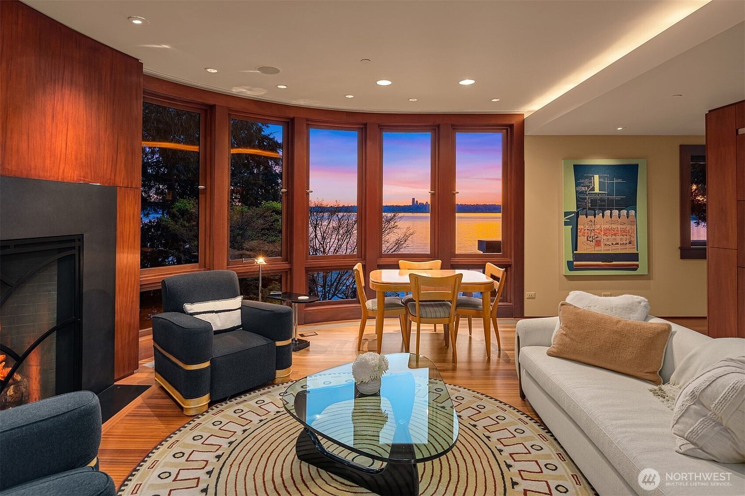 This elegant living room features a warm, contemporary design with rich wood paneling and a cozy fireplace. A large, curved window wall provides a stunning panoramic view of the lake at sunset, seamlessly blending indoor comfort with natural beauty. The space is furnished with plush seating, a unique glass-topped coffee table, and a dining area, creating an inviting atmosphere for relaxation and entertaining.
