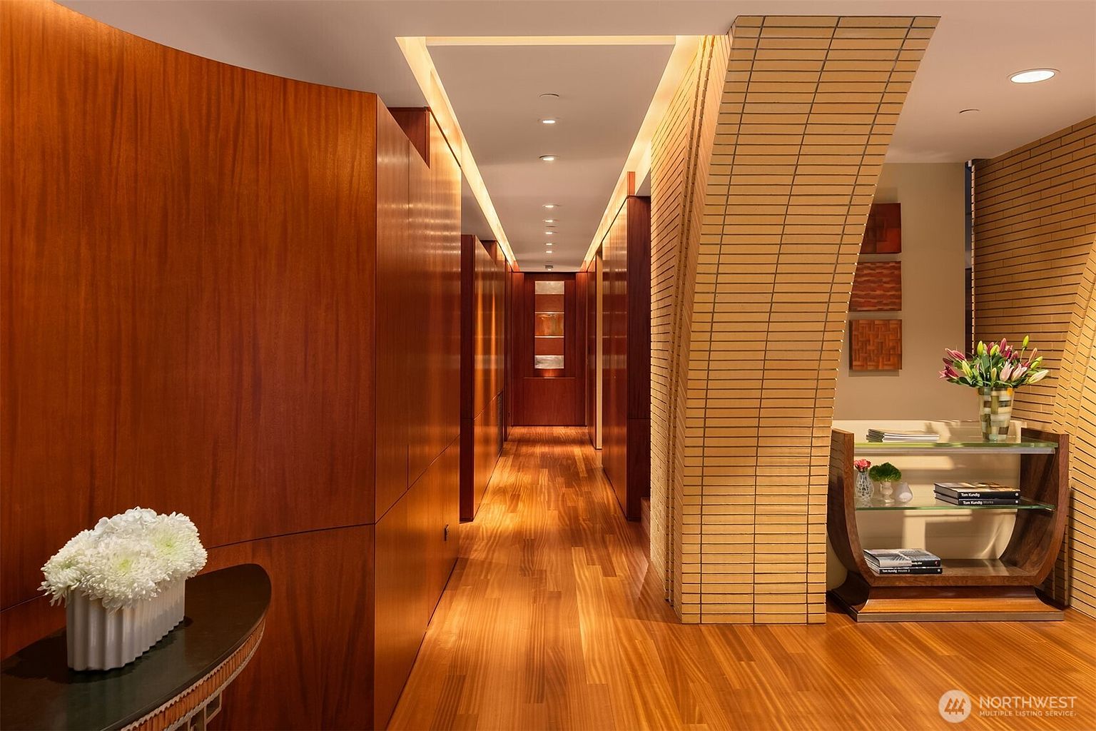 This sophisticated hallway features rich, warm-toned wood paneling and matching hardwood flooring, creating a seamless and luxurious aesthetic. A striking curved brick accent wall adds architectural texture, while recessed lighting guides the eye down the corridor toward a decorative glass-paneled door. The space is elegantly appointed with a sleek console table, a modern glass display shelf, and tasteful floral arrangements, evoking a high-end, contemporary atmosphere.
